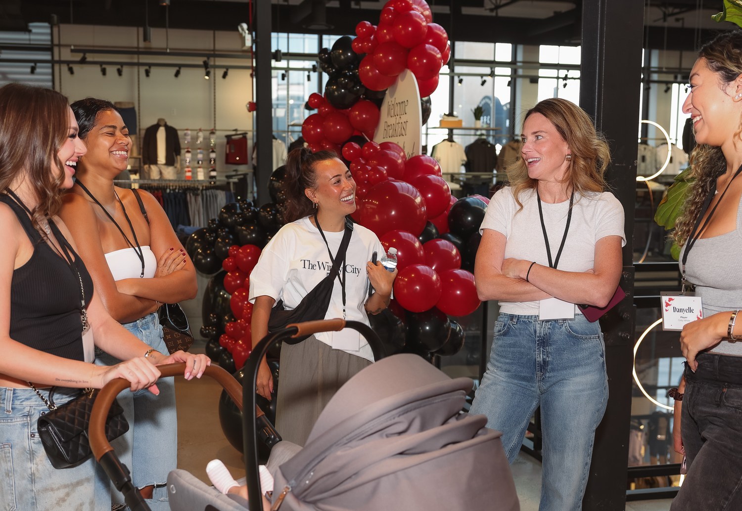 Partners talking together beside a red and black balloon arch at the welcome brunch, an infant in a stroller in the foreground